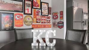 A dining area with a table, chairs, and a wall decorated with sports memorabilia and a large illuminated 'KC' sign. - Chief Themed 2 BR APT_KingBeds_Near Dining (Kansas City)