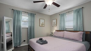 Une chambre à coucher comprenant un lit, un ventilateur de plafond, une armoire et deux fenêtres avec des rideaux.