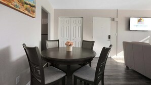 Un coin repas avec une table ronde, six chaises et un vase de fleurs. Un téléviseur est fixé au mur, et une porte est visible au fond.