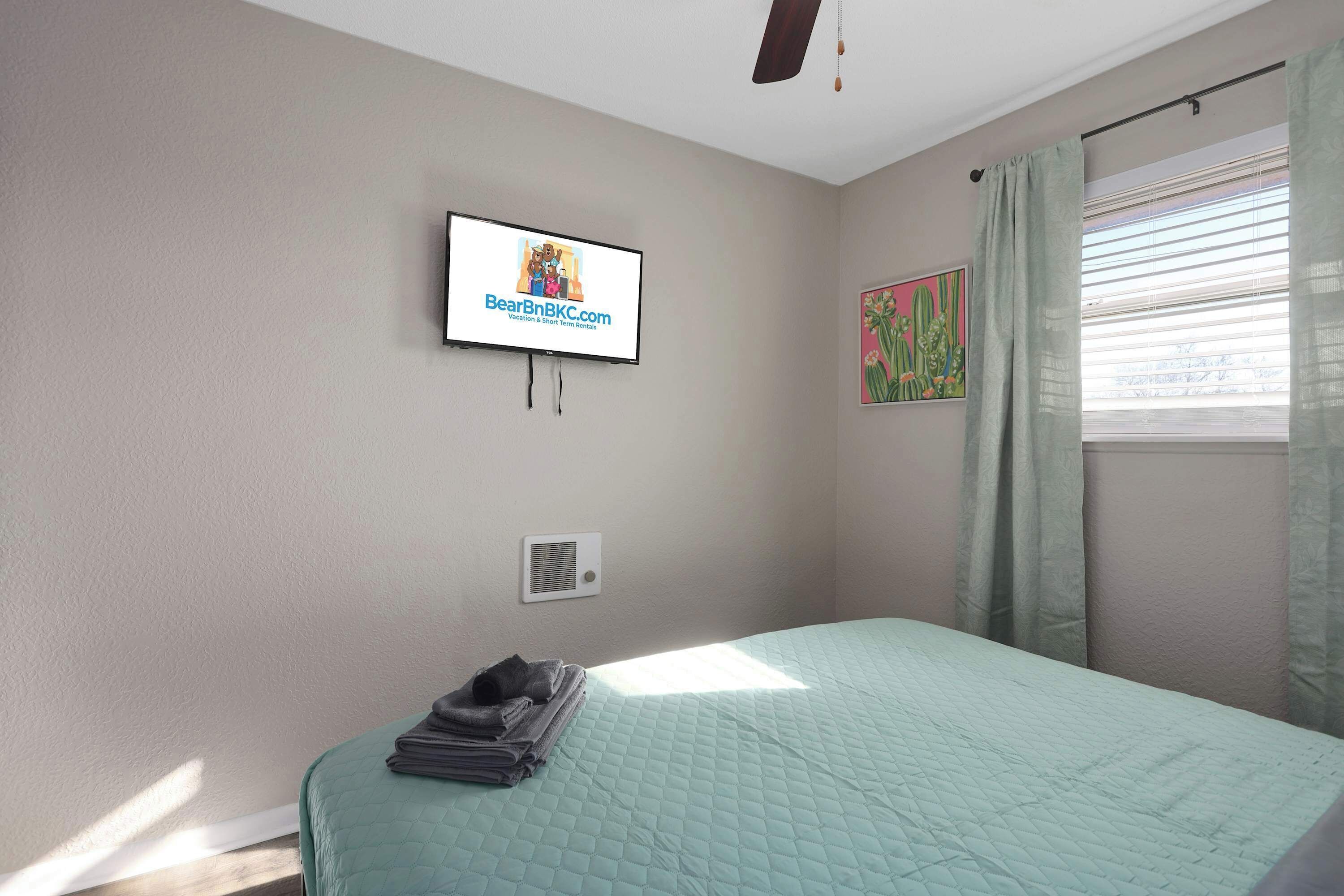 A bedroom with a bed, a TV mounted on the wall, a window with curtains, and a wall-mounted air conditioning unit.