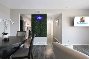 Ein modernes Wohnzimmer mit Essbereich, einer grünen Wand mit einer Neonreklame und einem Fernseher, der eine Website für Ferienwohnungen zeigt.