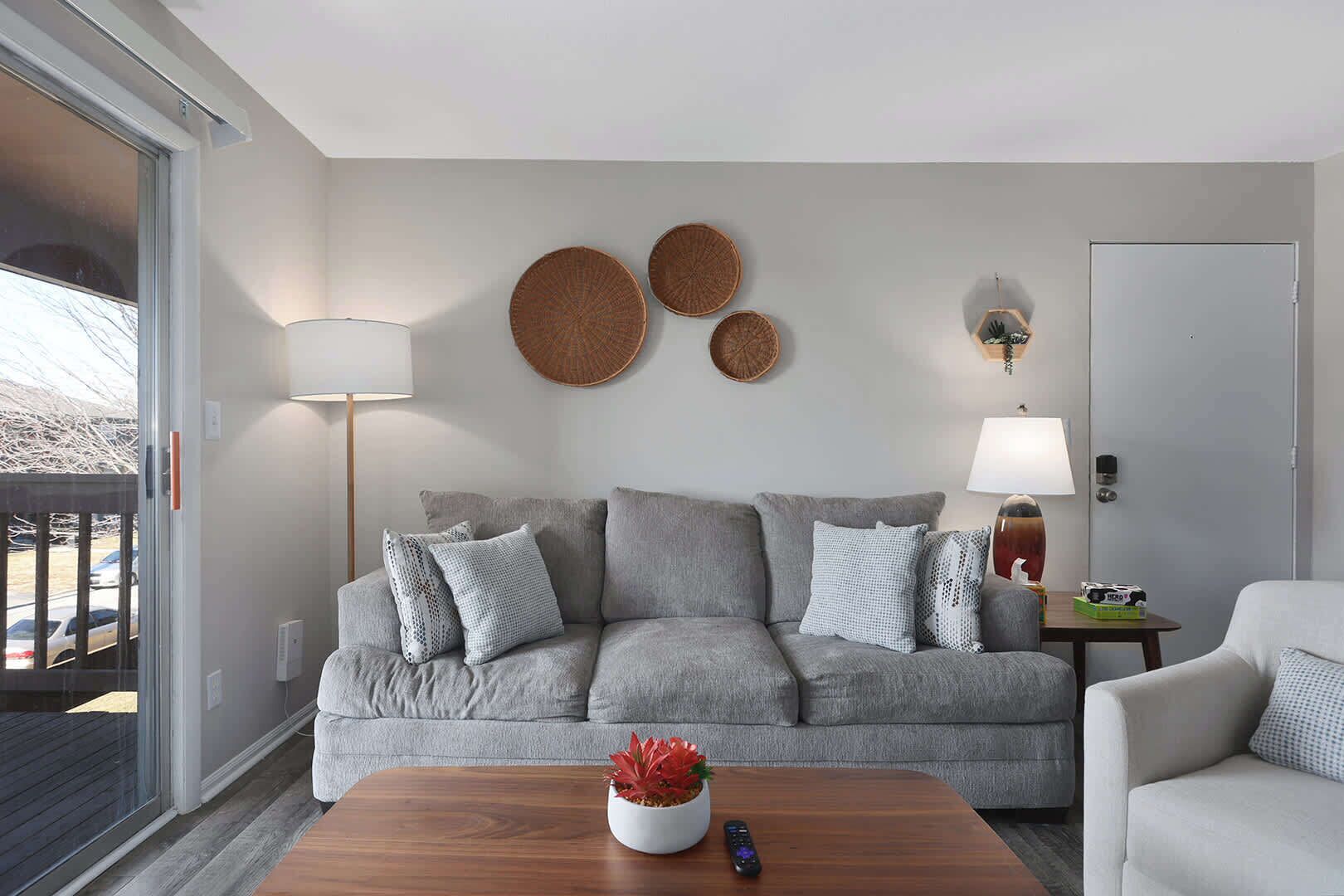 A living room with a grey sofa, a wooden coffee table with a plant, and a white armchair. There are two wall-mounted lamps and a door with a handle.
