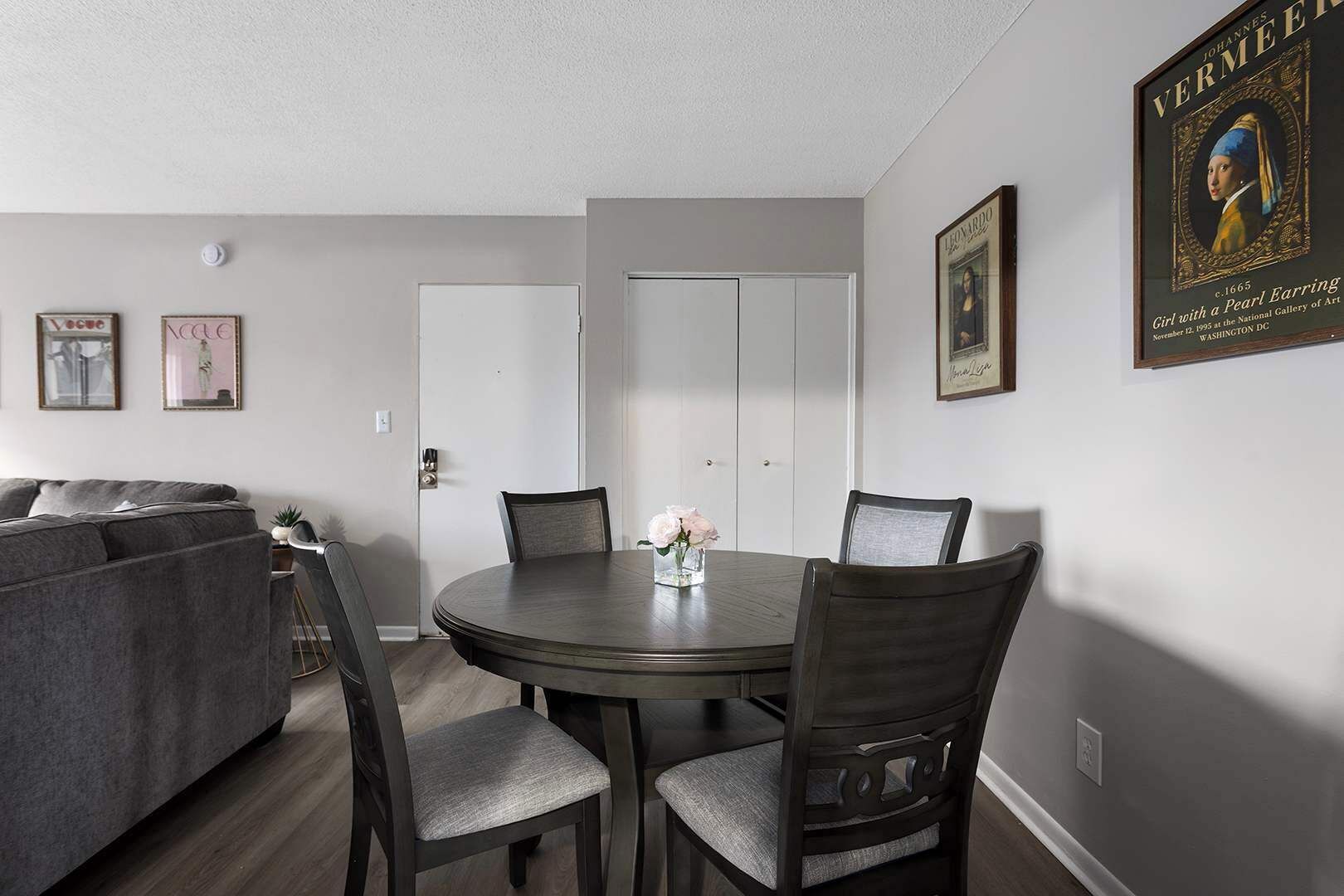 A dining area with a round table, four chairs, and a vase with flowers. There is a couch in the background and a door to the right.