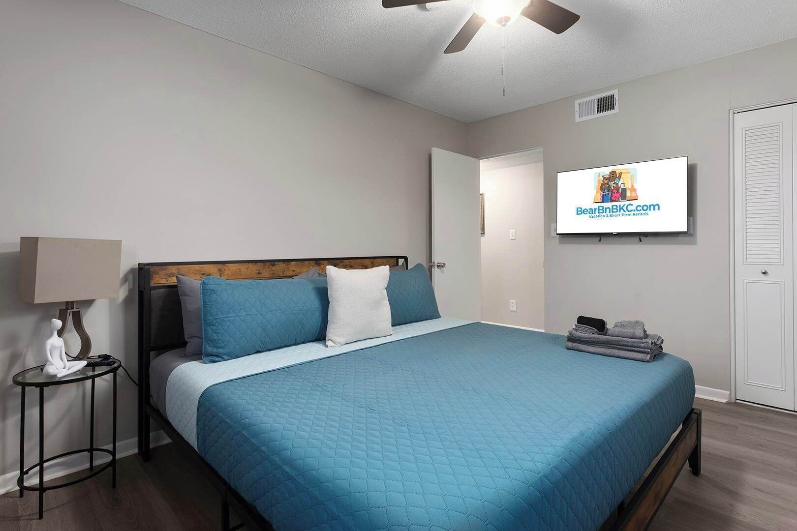 A bedroom with a bed, a nightstand, a lamp, a ceiling fan, a TV, and a door.
