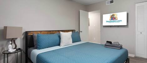 A bedroom with a bed, a nightstand, a lamp, a ceiling fan, a TV, and a door.