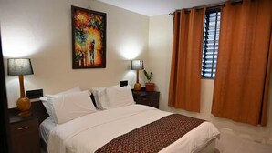 A hotel room with a bed, two bedside tables, a painting on the wall, a window with curtains, and a striped rug. - Lavish Lodge (Suva)