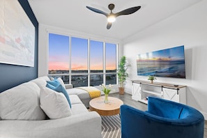 A modern living room with a large window, a ceiling fan, a sofa, a blue armchair, and a TV on a wooden stand. - Sunset Penthouse: Pool, Spa, Gym, Games, & Parking (San Diego)