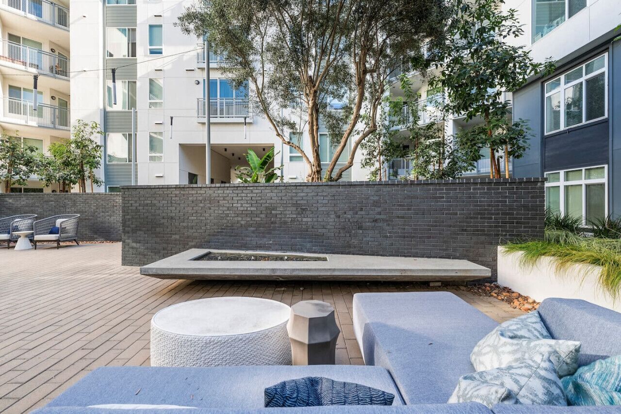 Un moderno espacio lounge al aire libre con asientos y una chimenea, rodeado por un muro de ladrillo y follaje.