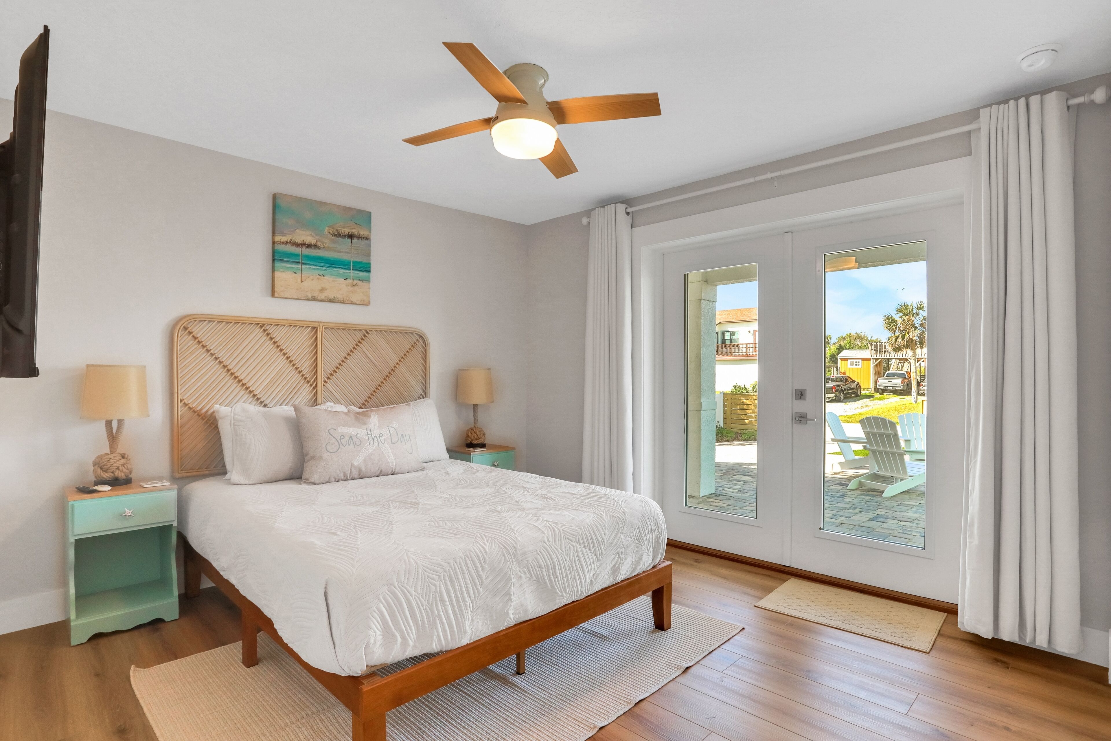 Un dormitorio con cama de madera, ventilador de techo, un cuadro con motivos playeros y vistas al exterior a través de una puerta corrediza.
