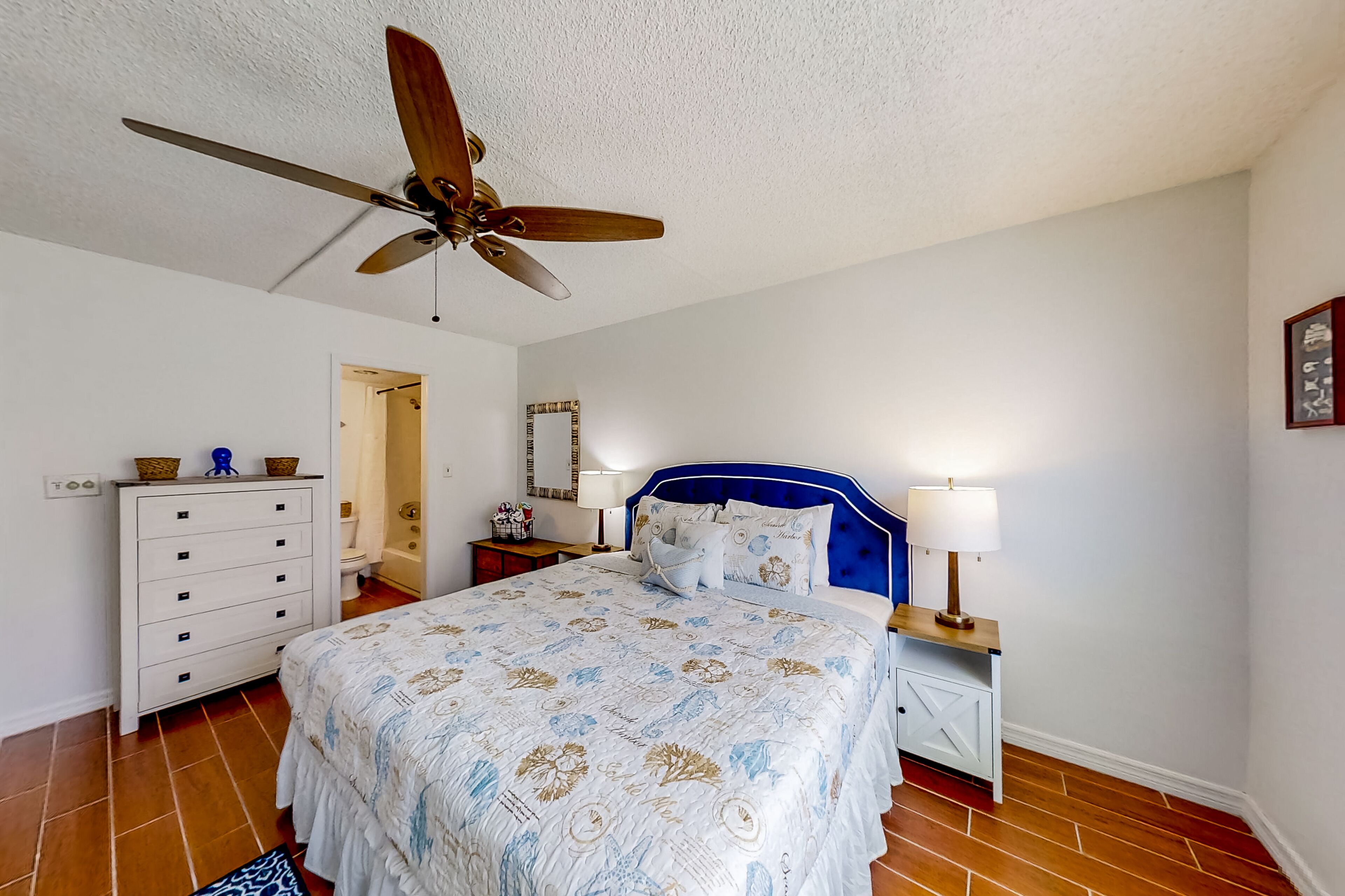A bedroom with a bed, a ceiling fan, a dresser, a nightstand, and a lamp.