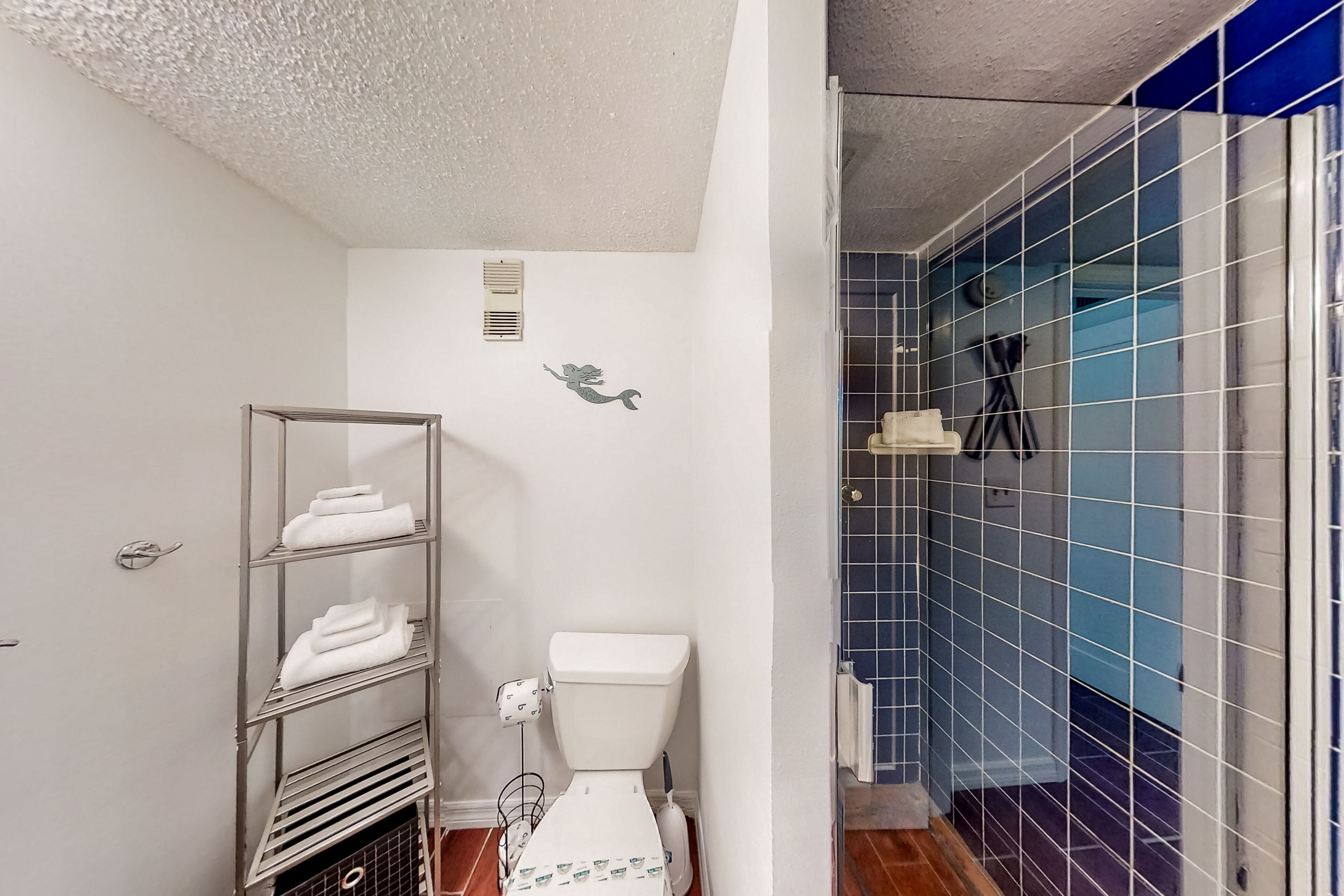 A bathroom with a white toilet, a metal towel rack with folded towels, a wall-mounted vent, and a shower area with blue-tiled walls.