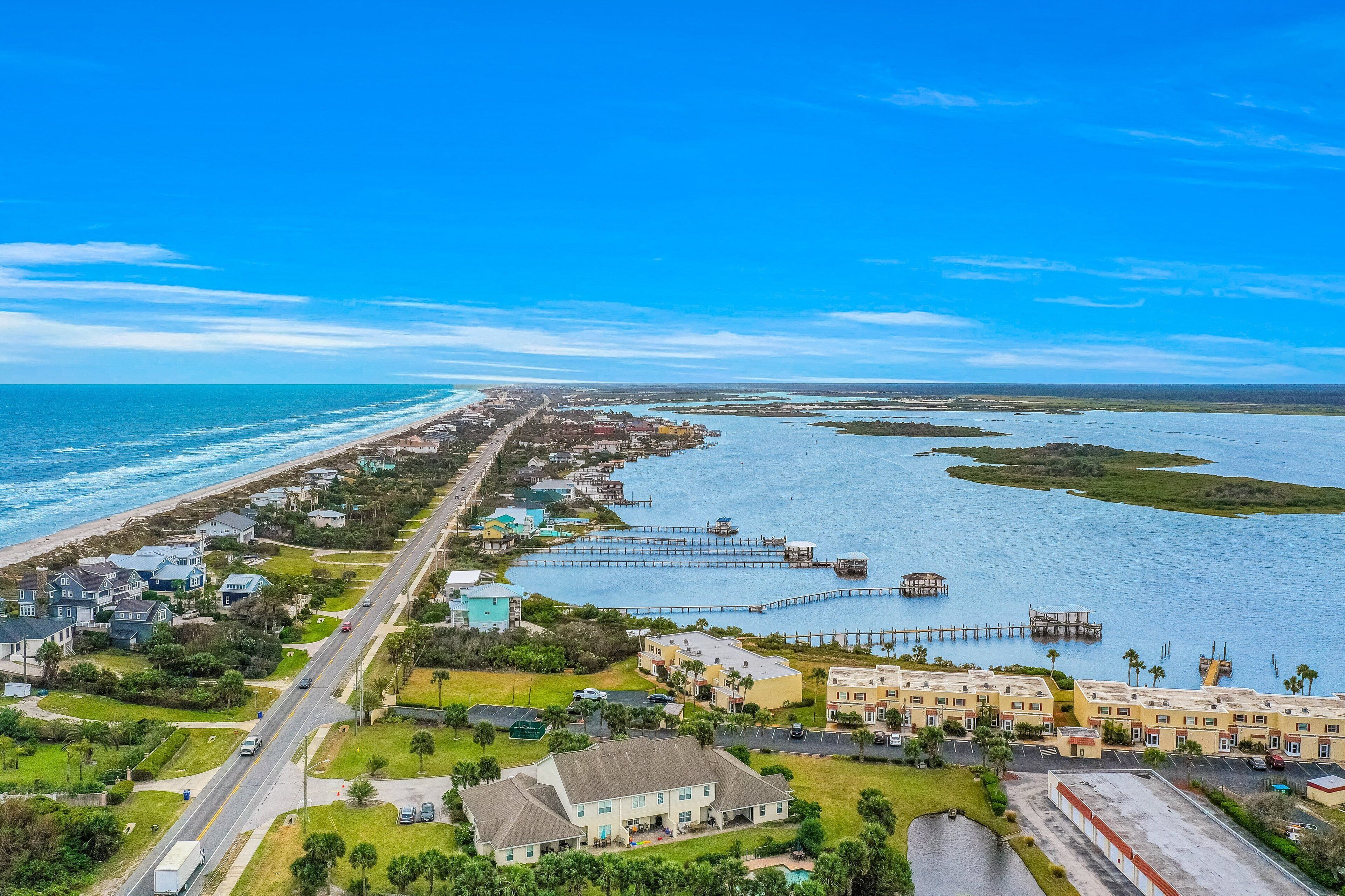 An aerial view of a coastal area with a long road, beachfront properties, and a marina with docks.