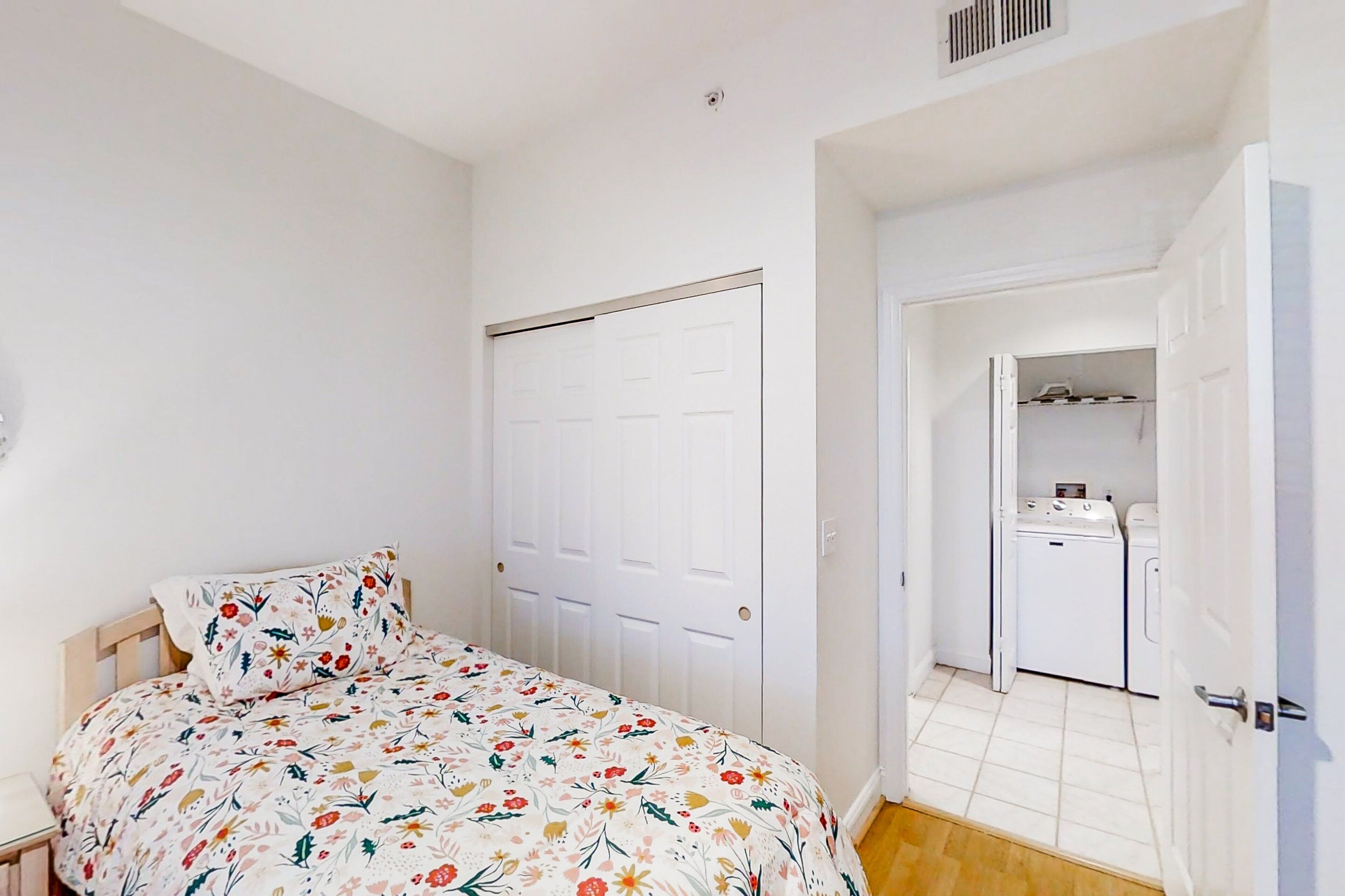 A bedroom with a bed, a white door, and a small kitchenette in the background.