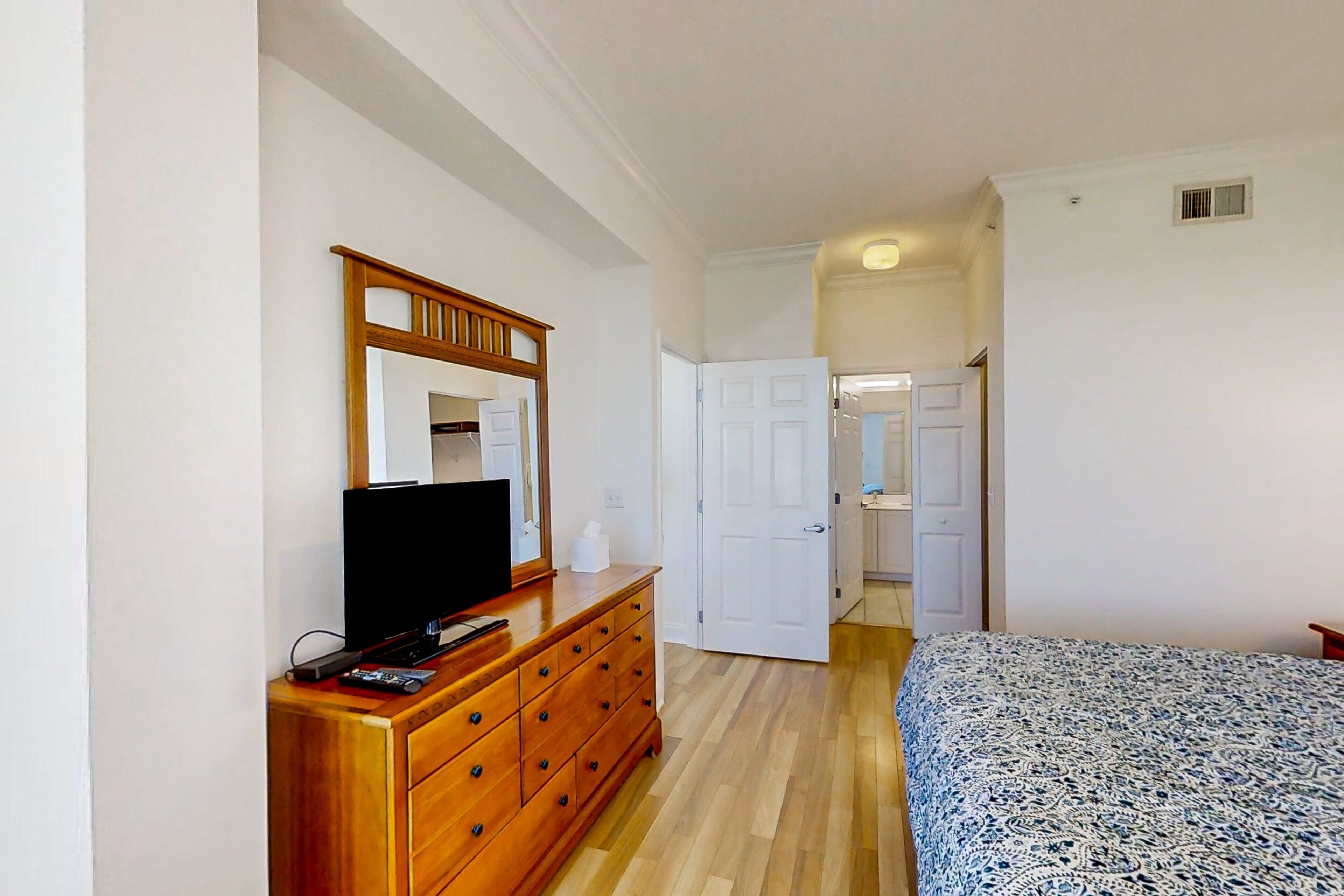 A bedroom with a wooden dresser, a television, a bed with a patterned bedspread, and a door leading to another room.