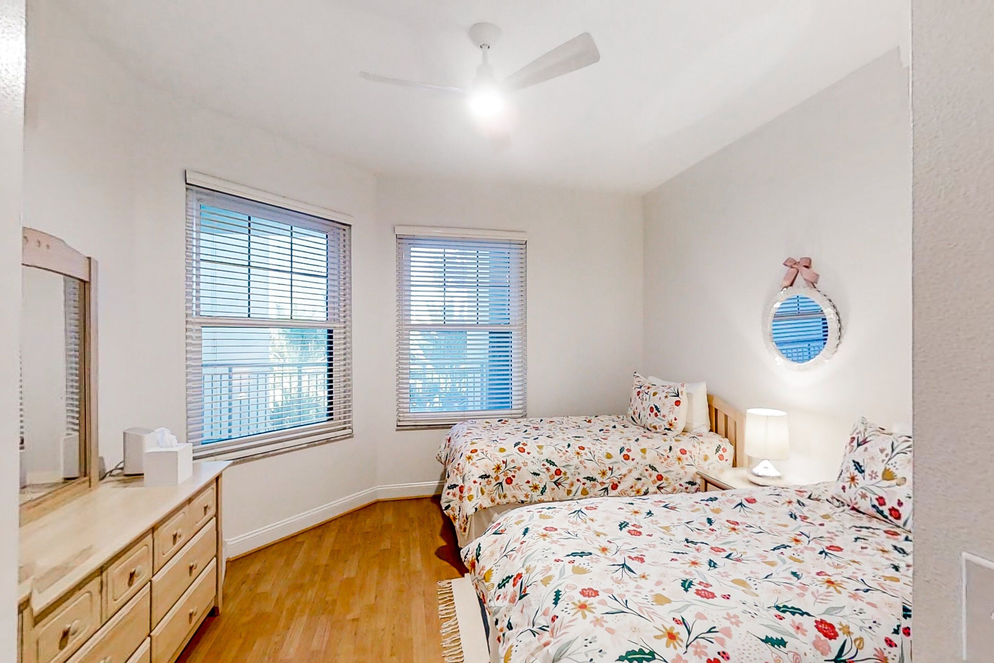 A bedroom with two beds, a dresser, a ceiling fan, and two windows with blinds.