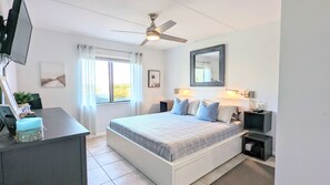 A bedroom with a bed, bedside tables, a ceiling fan, a window with curtains, a wall-mounted TV, and a mirror. - Cool Waves Oceanfront Retreat - St Augustine Beach 2 Pools 5 Hot Tubs! On the Beach (St. Augustine)