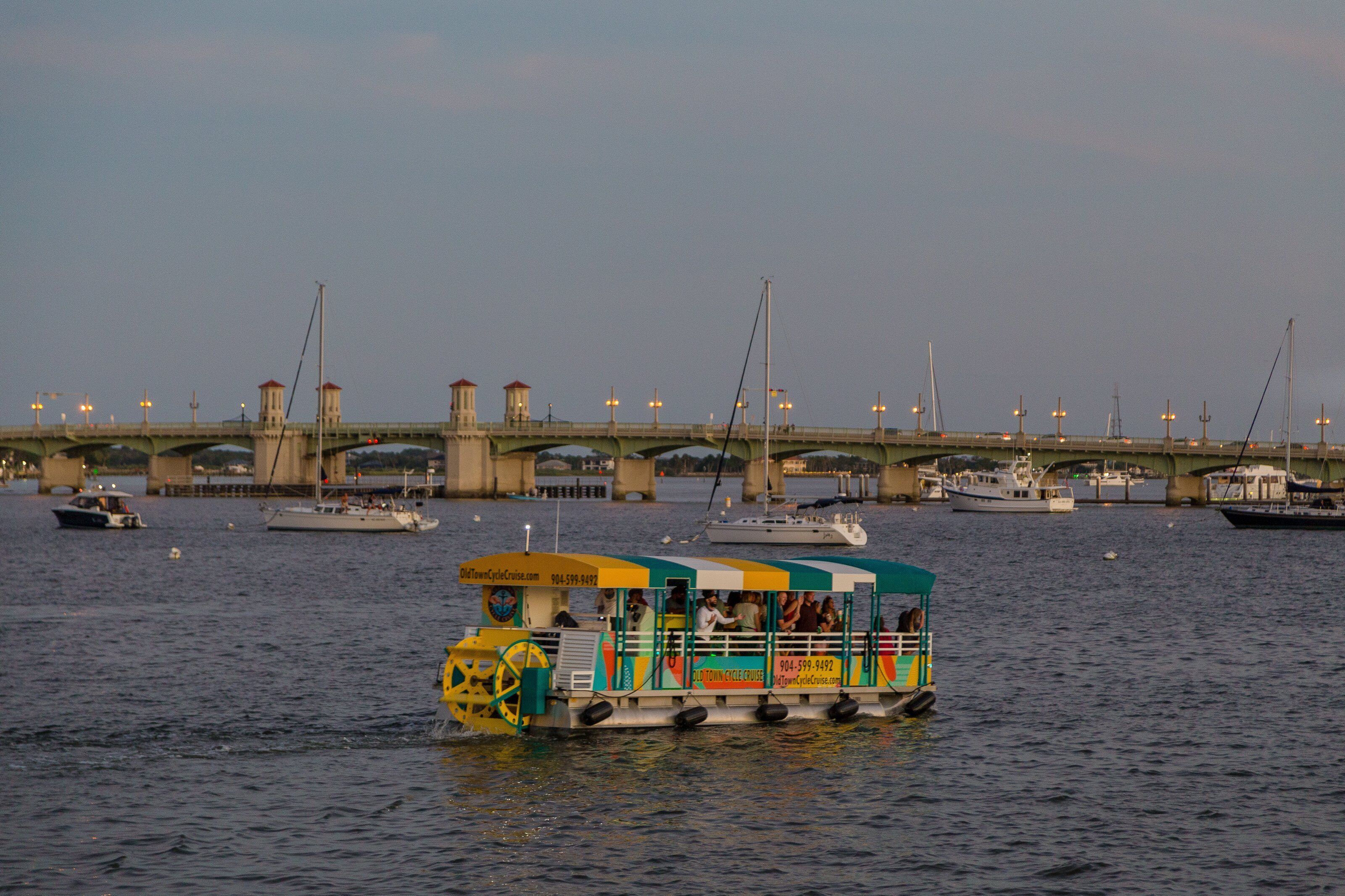 Ein buntes Boot mit einer gelben Fahrkabine und grünen und gelben Akzenten, das auf einem Fluss treibt, im Hintergrund ist eine Brücke zu sehen.