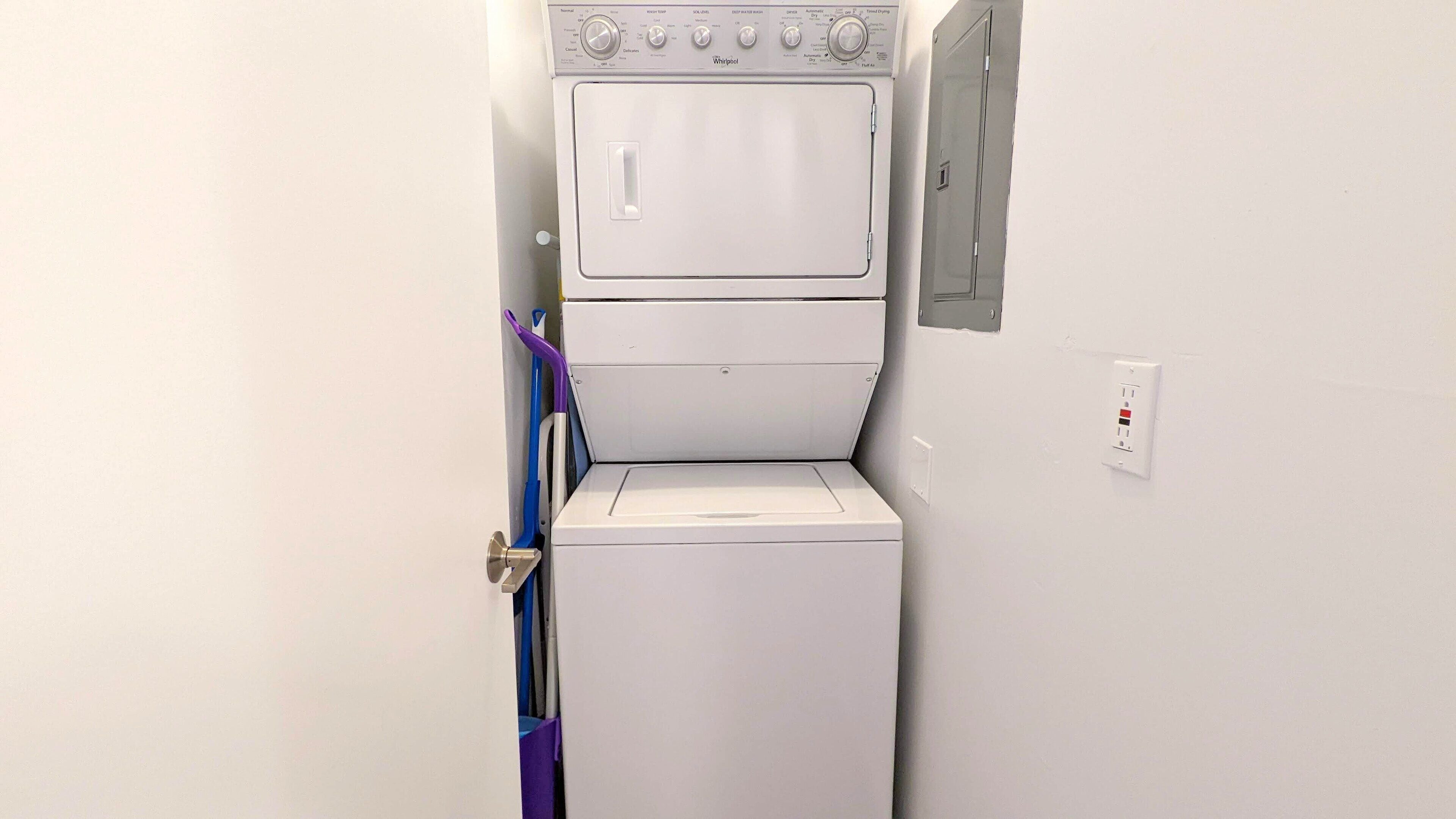 Une petite buanderie blanche, équipée d’un lave-linge et d’un sèche-linge à chargement frontal. Un balai violet est appuyé contre le mur.