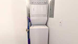 A compact, white laundry room with a front-loading washer and dryer. There is a purple mop leaning against the wall. - Cool Waves Oceanfront Retreat - St Augustine Beach 2 Pools 5 Hot Tubs! On the Beach (St. Augustine)