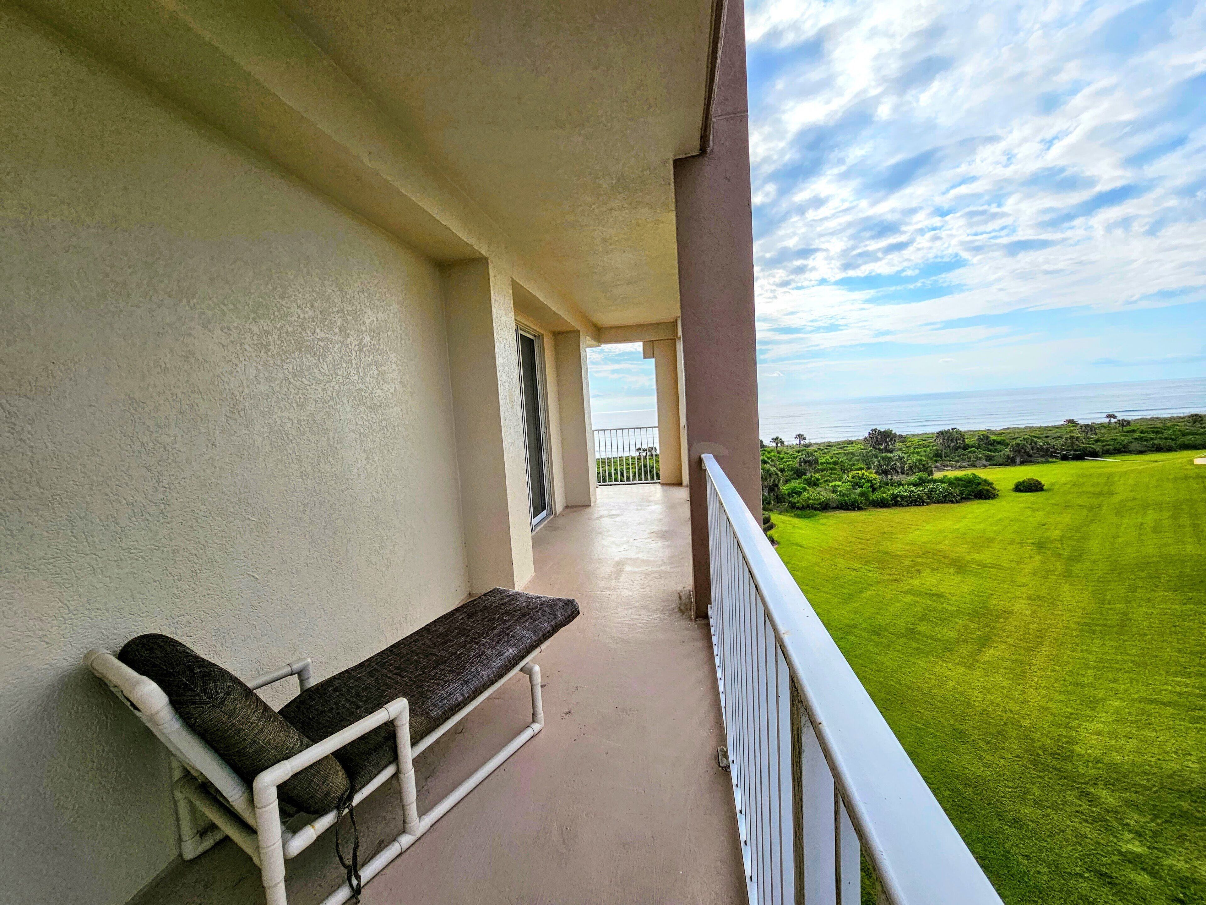 A balcony with a white railing, a bench, and a view of a green lawn and ocean.