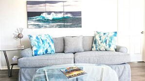 Ein Wohnzimmer mit einem grauen Sofa, zwei blau-weißen Kissen mit Muster, einem Glastisch mit einer Zeitschrift und einem Wandbild einer Welle.