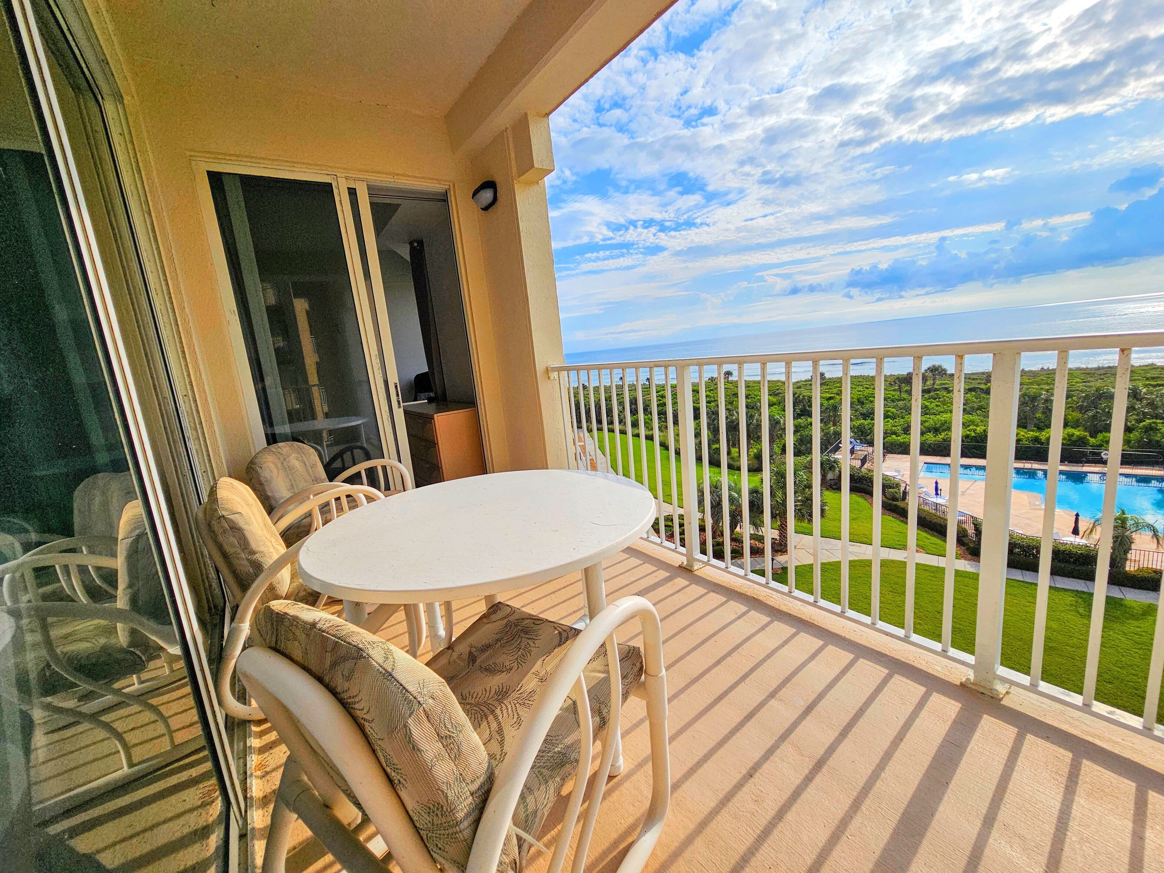 A balcony with a white table and chairs, overlooking a pool and lush greenery.