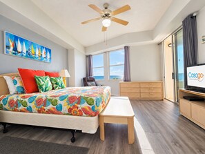 A hotel room with a bed, a desk, a television, a ceiling fan, a window with a view, and a painting on the wall. - Breathtaking 5th Floor Ocean Views. Corner Unit Wraparound Balcony! 3 Pools (Palm Coast)