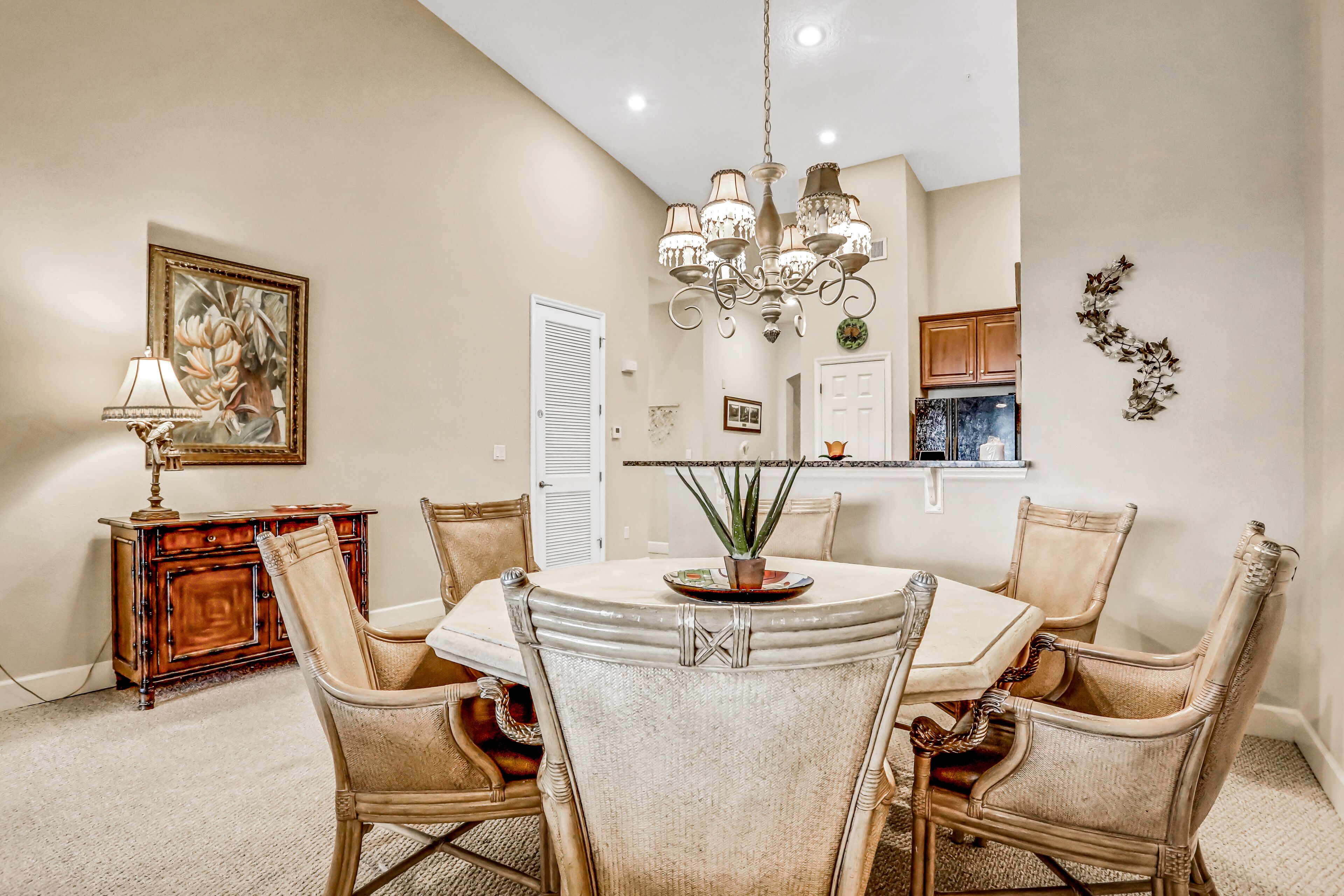 A dining area with a round table, six chairs, a chandelier, a painting, and a cabinet.