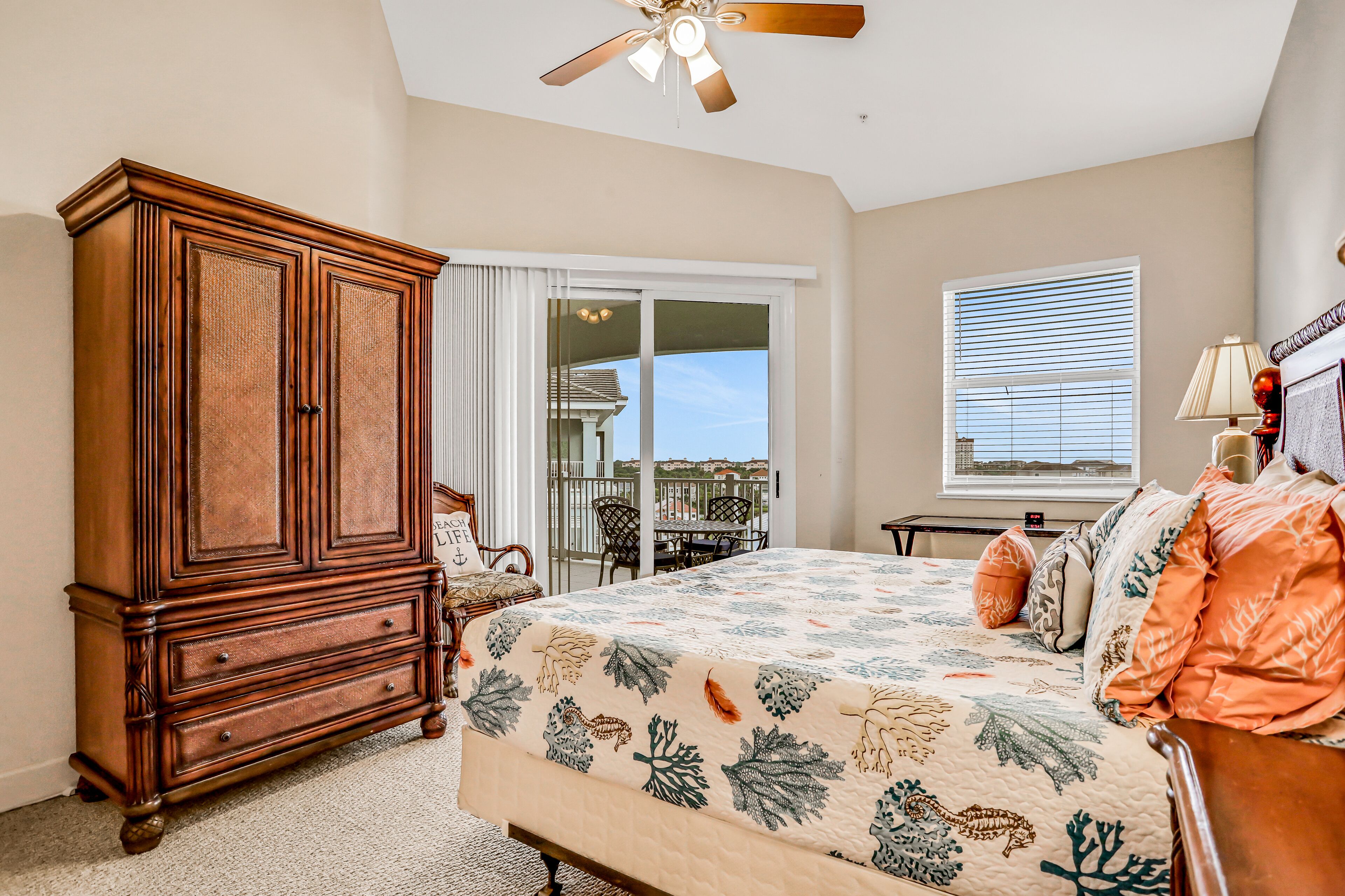 A bedroom with a large wooden wardrobe, a bed with a floral bedspread, a balcony with a table and chairs, and a window with blinds.