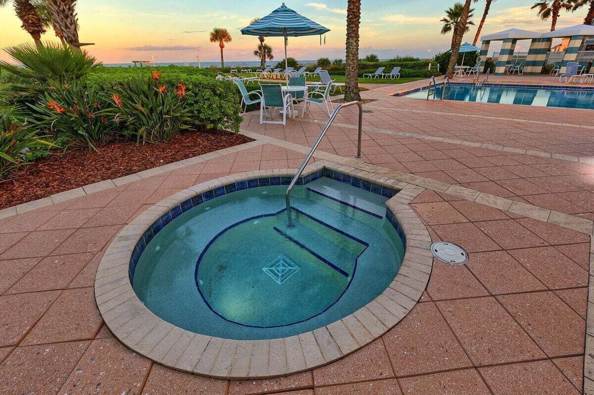 A small, circular hot tub surrounded by a tiled patio with lounge chairs and a pool in the background.