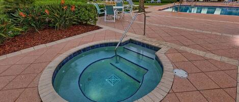 A small, circular hot tub surrounded by a tiled patio with lounge chairs and a pool in the background.