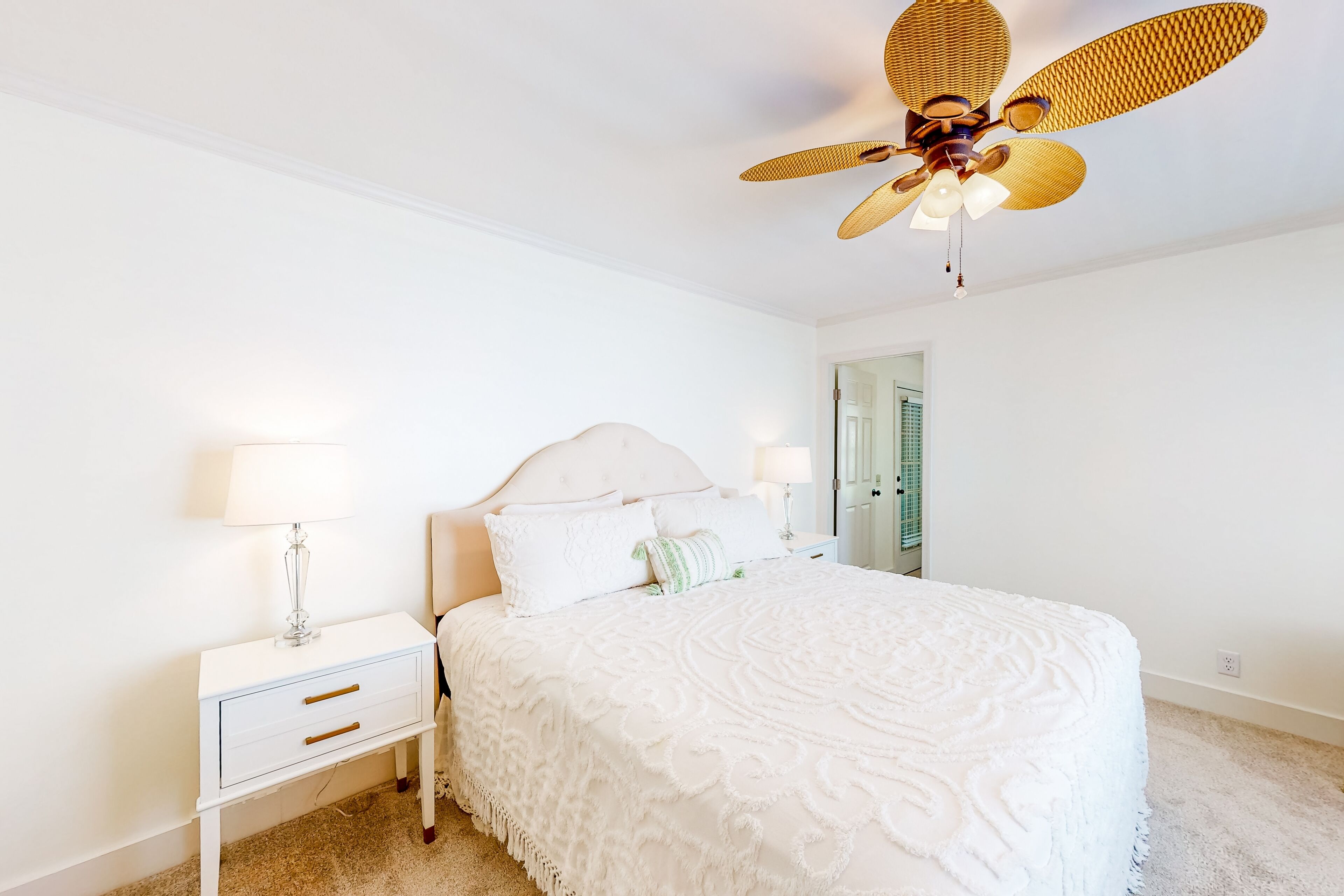 Un dormitorio con una cama grande, paredes blancas, ventilador de techo y una mesita de noche con lámpara.