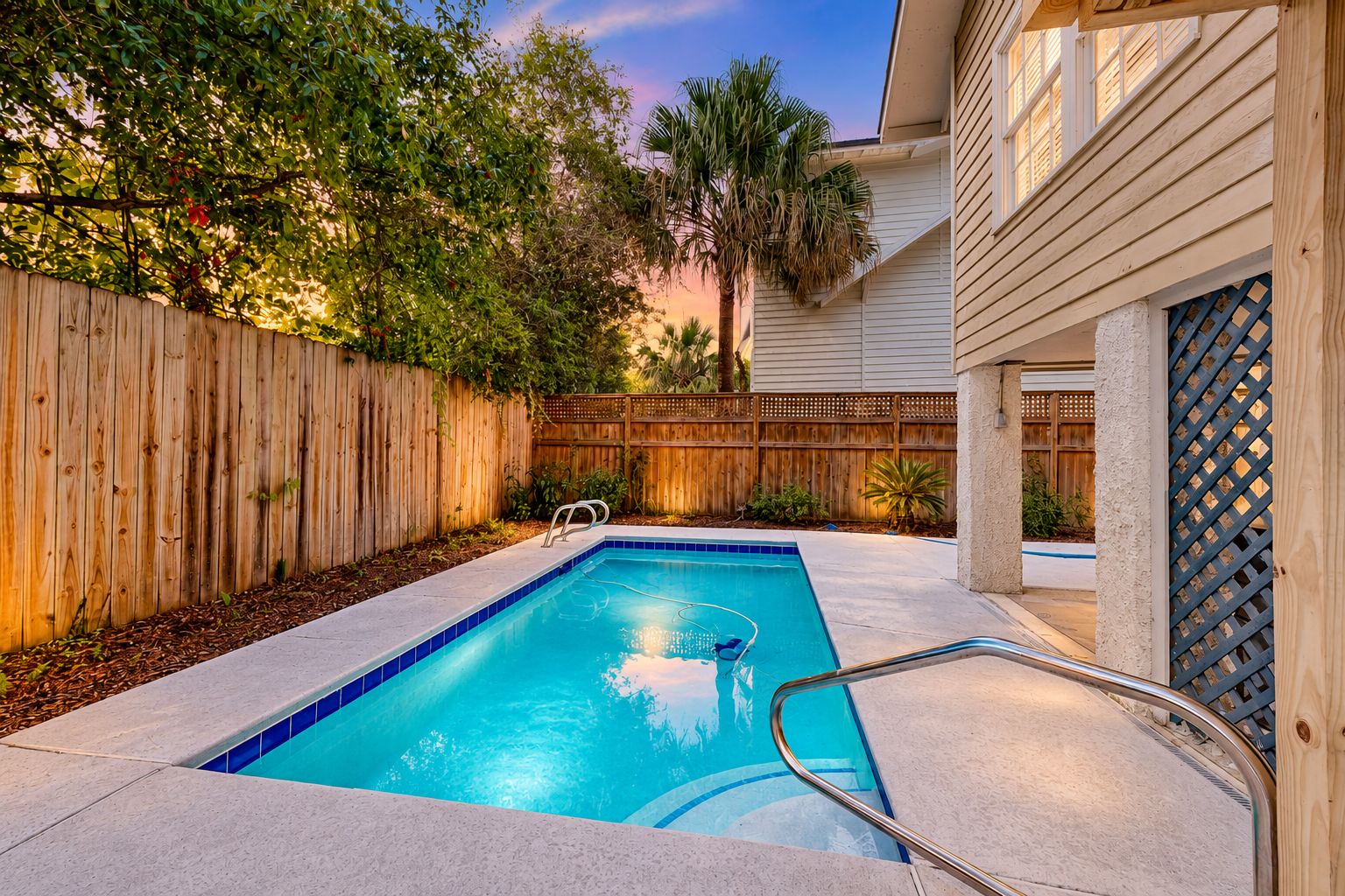 A rectangular swimming pool with a metal ladder, surrounded by a wooden fence and various plants.