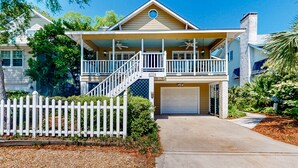A two-story house with a front porch, a garage, and a white picket fence. - Classic Coastal Home With Game Room & Private hot tub - Walk to Beaches (St. Simons Island)
