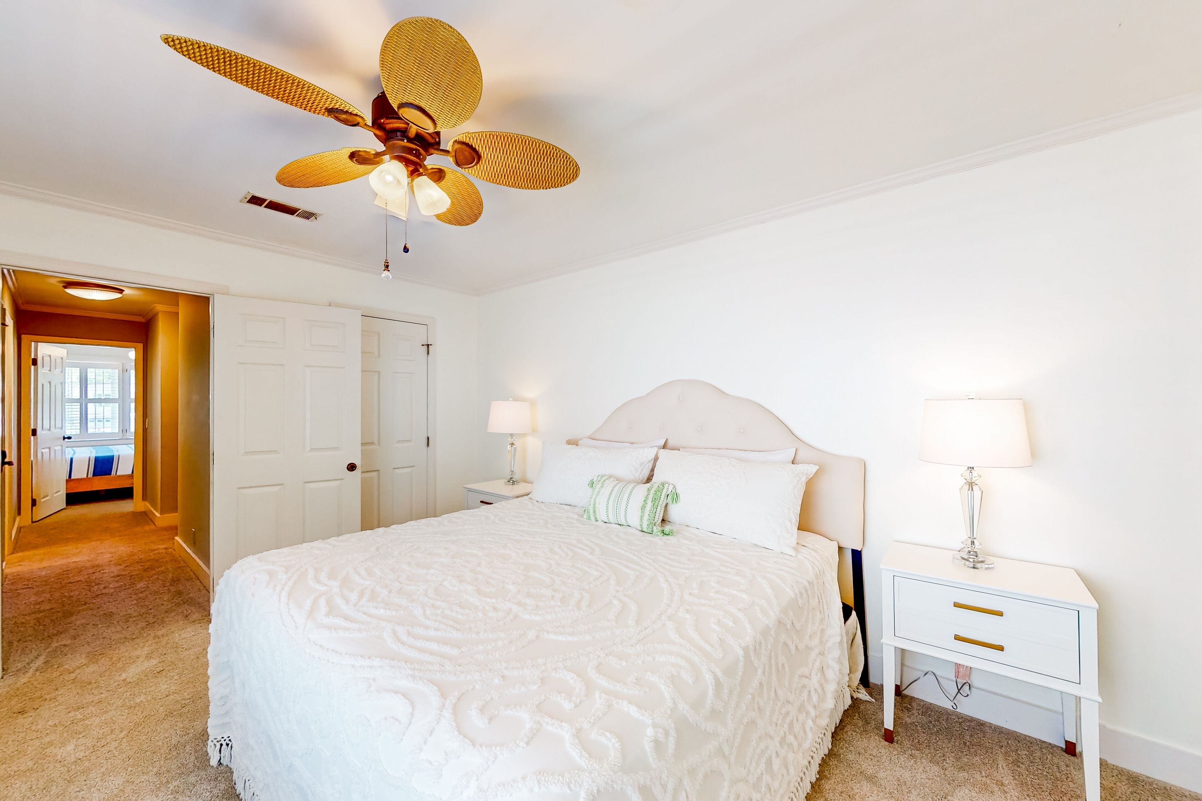 A bedroom with a large bed, a ceiling fan, a nightstand with a lamp, and a door leading to another room.