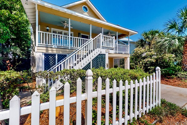 A two-story house with a porch, a white picket fence, and a well-maintained garden. - Classic Coastal Home With Game Room & Private hot tub - Walk to Beaches (St. Simons Island)