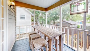 A covered porch with a wooden table and two cushioned chairs, a white railing, and a stained glass window. - Classic Coastal Home With Game Room & Private hot tub - Walk to Beaches (St. Simons Island)