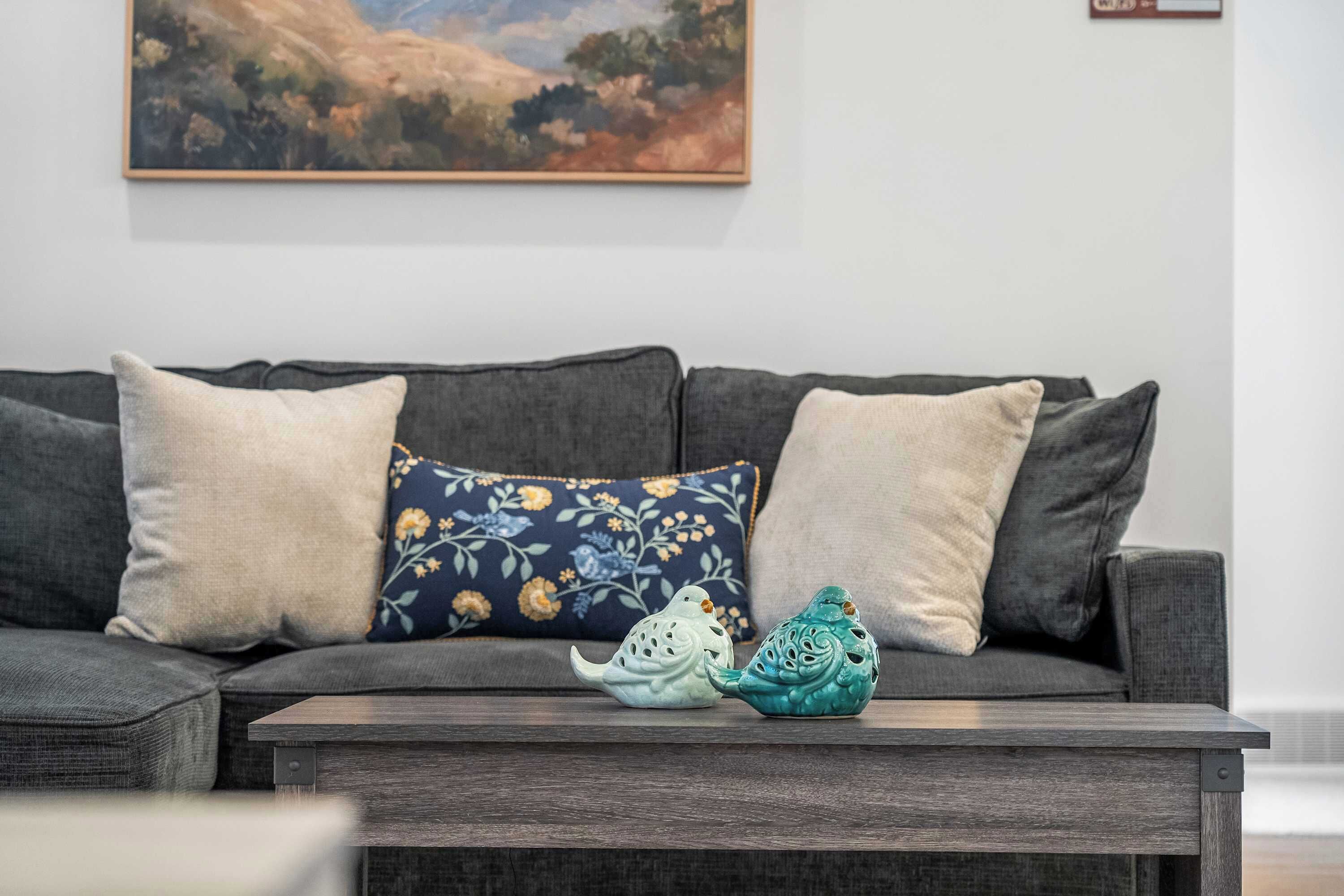 Una sala de estar moderna con un sofá gris, dos almohadones decorativos y una mesita con dos jarrones con forma de pájaro.