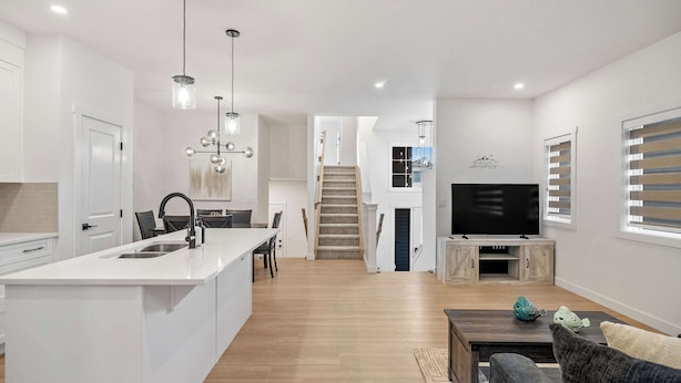 A modern kitchen with a central island, a dining area with chairs, a staircase leading to an upper level, and a living room with a TV and decorative items. - 5BR Family Home w Kids Play Den (Calgary)