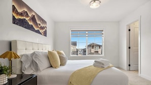 A bedroom with a large bed, a bedside table with a lamp, a window with a view of buildings, and a painting on the wall. - 5BR Family Home w Kids Play Den (Calgary)