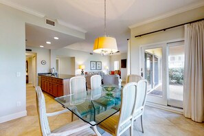 A dining area with a glass table and chairs, a kitchen in the background, and a sliding glass door leading to an outdoor area. - 1801 Turtle Dunes (Fernandina Beach)