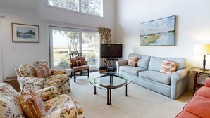A spacious living room with a large window, a flat-screen TV, a glass coffee table, and a light blue sofa. - Amelia Island Plantation Townhome With Amazing Marsh Views, Wifi & Shared Pool (Fernandina Beach)
