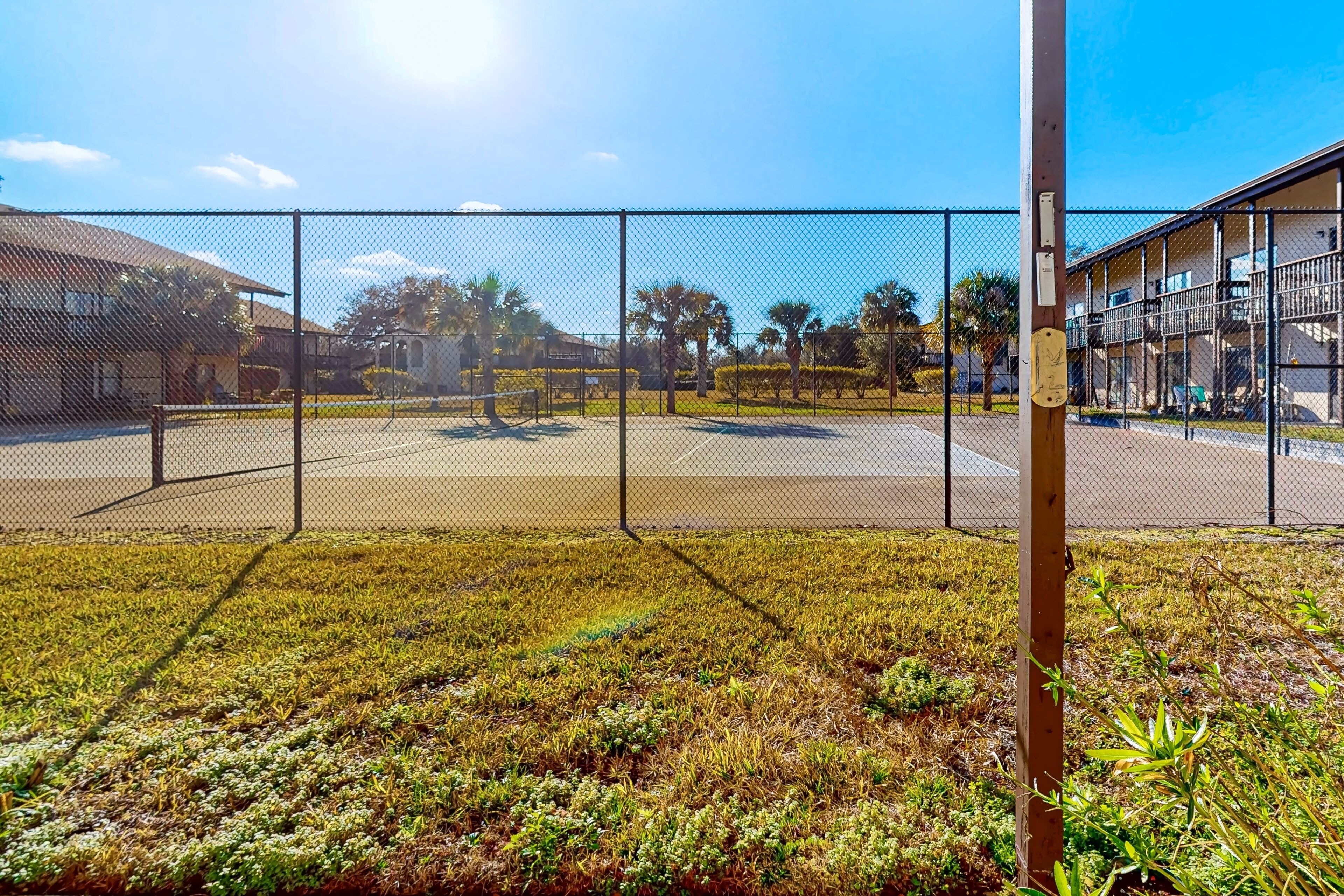 Uma área externa cercada com quadra de basquete, um edifício com varanda e céu claro.