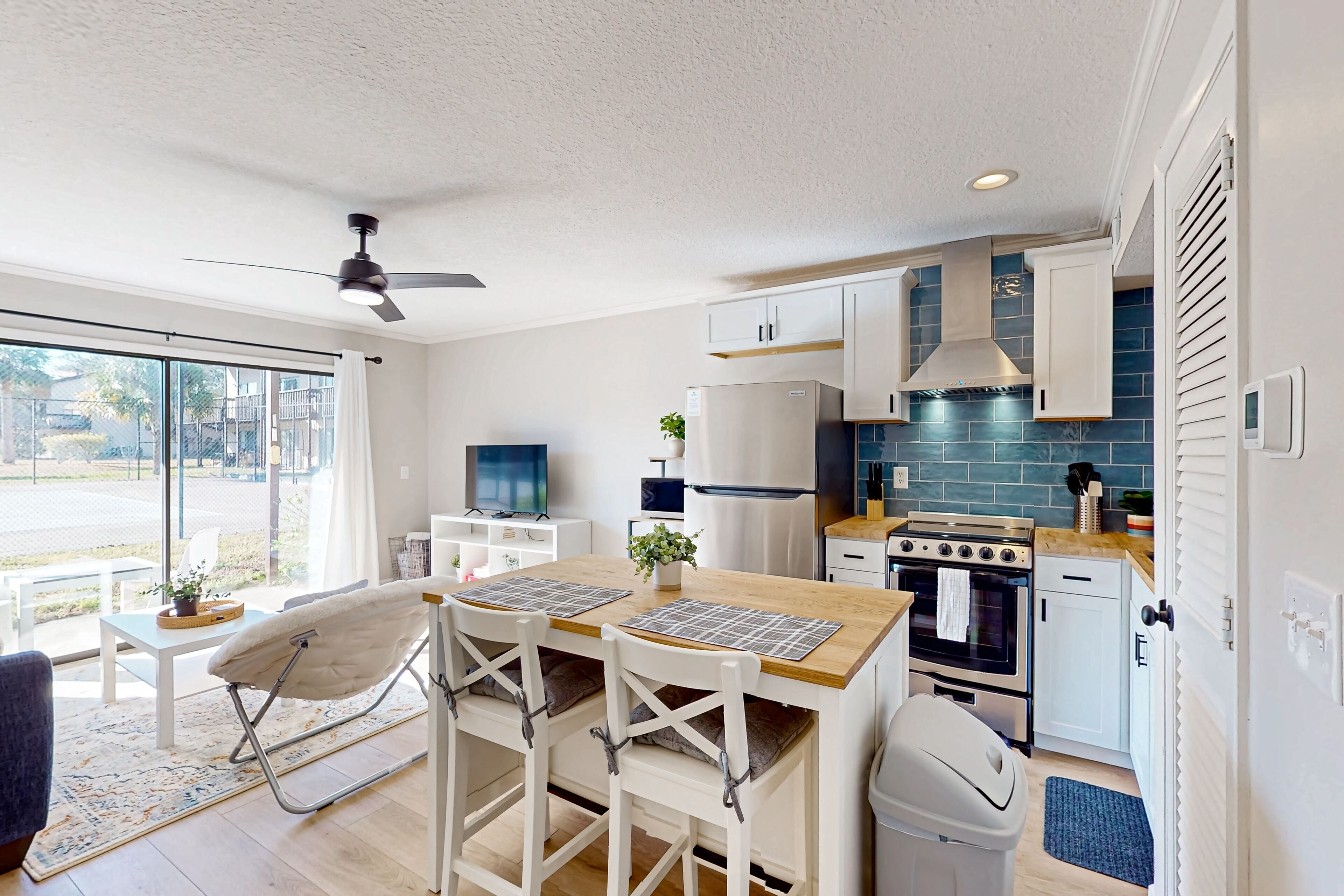 A modern kitchen with white cabinets, a wooden countertop, and a built-in oven. There is a dining area with a table and chairs, and a ceiling fan.