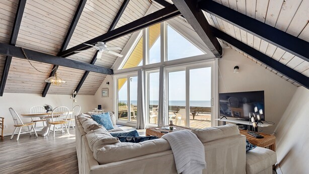 A spacious living area with a high ceiling, large windows, a sofa, a dining table, and a television. - A-frame Beach House Steps From the Ocean With Deck & Central AC (Ponte Vedra Beach)