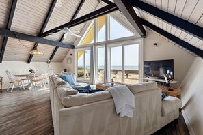 A spacious living area with a high ceiling, large windows, a sofa, a dining table, and a television. - A-frame Beach House Steps From the Ocean With Deck & Central AC (Ponte Vedra Beach)