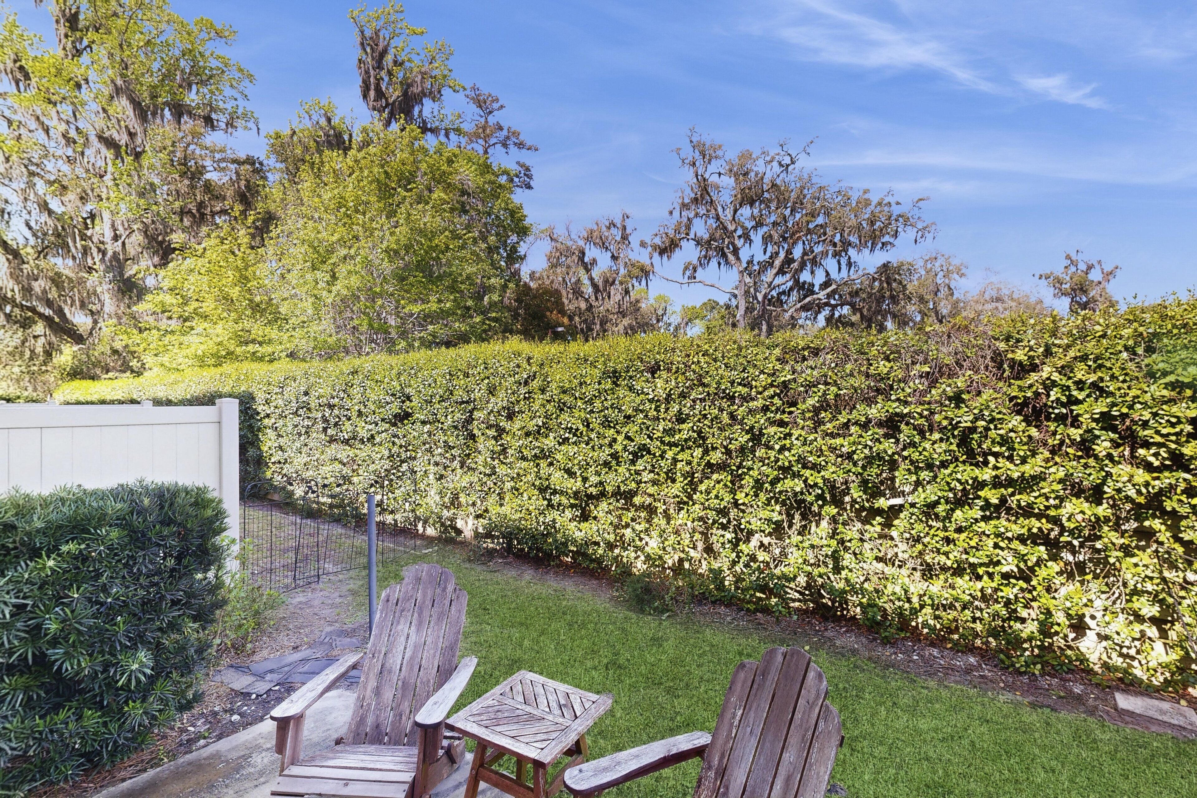 Un jardín con sillas y una mesa de madera, rodeado por un seto y árboles.