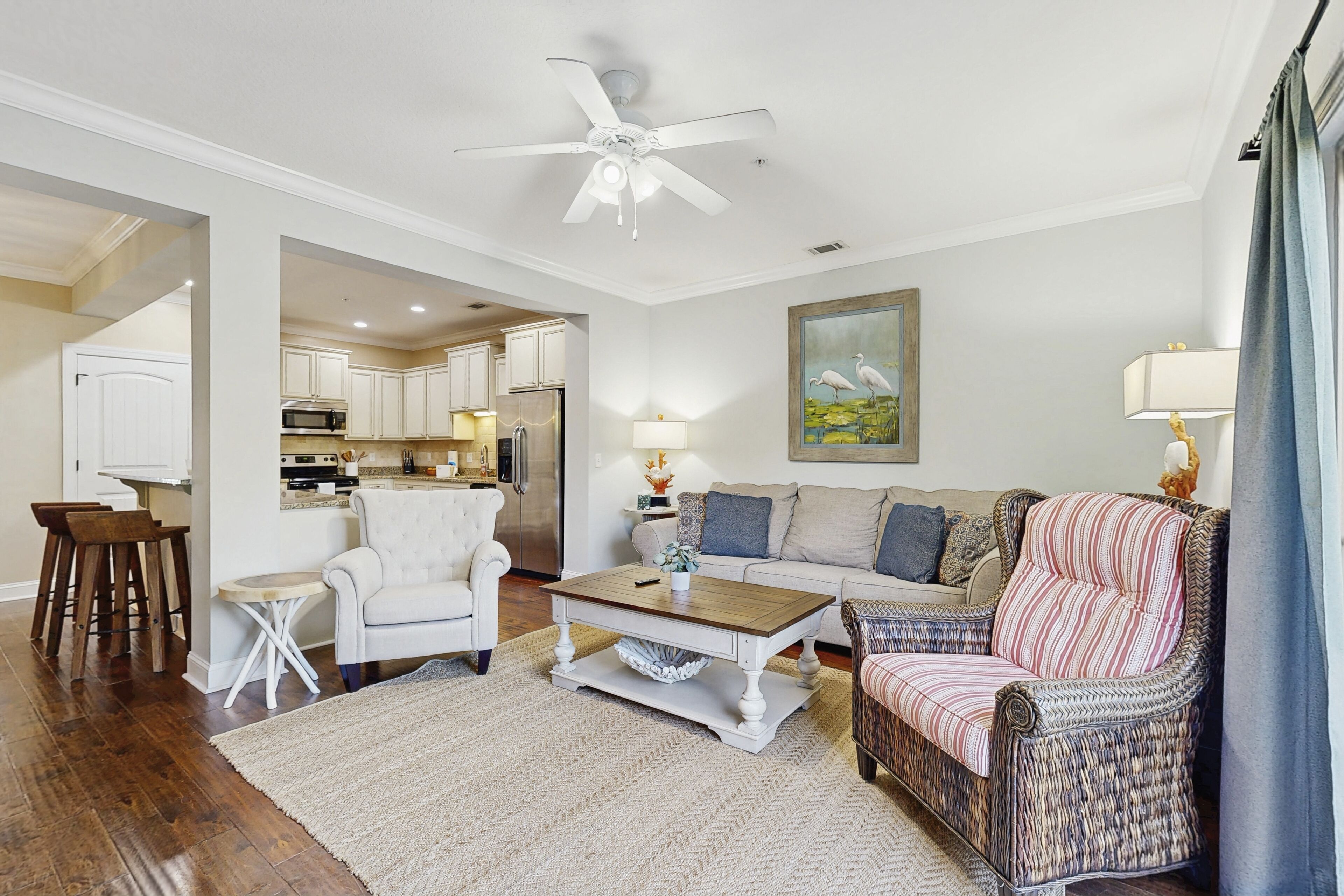 A spacious living room with a white ceiling fan, a dining area with wooden chairs, a kitchen with white cabinets, and a painting of birds on the wall.