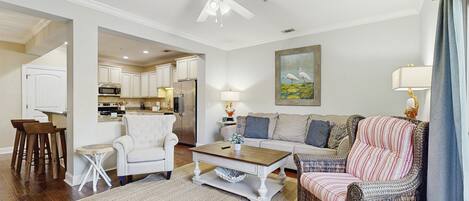 A spacious living room with a white ceiling fan, a dining area with wooden chairs, a kitchen with white cabinets, and a painting of birds on the wall.