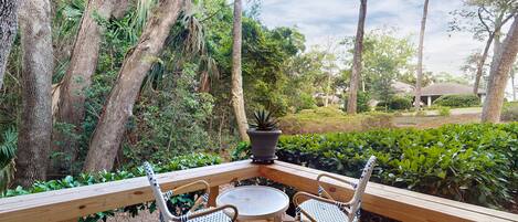A wooden deck with two wicker chairs and a table, surrounded by lush greenery and trees.